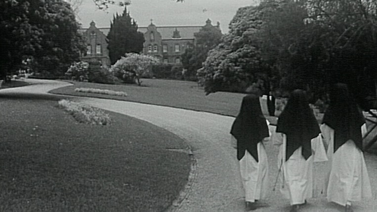 A black and white still from a video as 3 nuns walk up a garden path towards the convent.