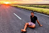a young man in casual clothes sits on a quiet road amid beautiful fields as the sun sets