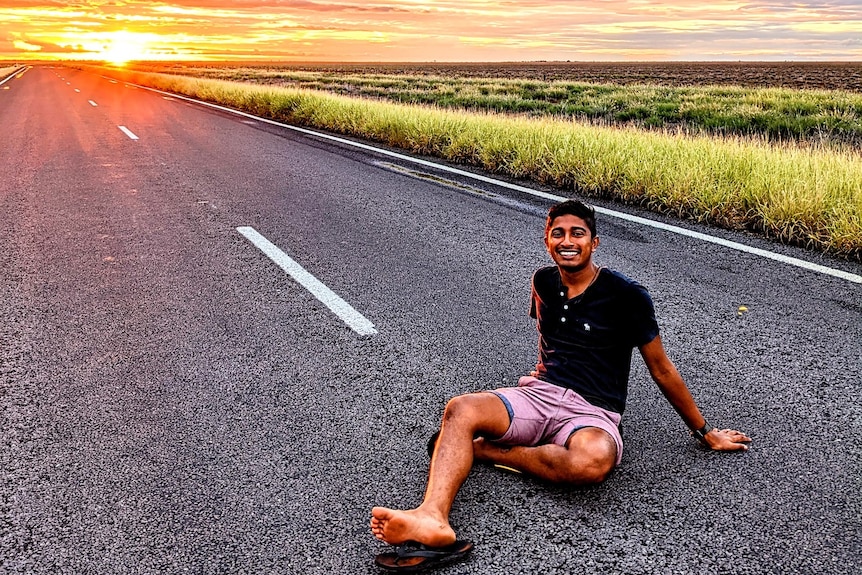 a young man in casual clothes sits on a quiet road amid beautiful fields as the sun sets