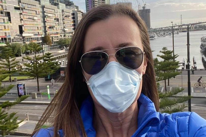 A selfie shot of WA nurse Renee Freeman wearing a blue jacket, face mask and sunglasses in front of a city backdrop.