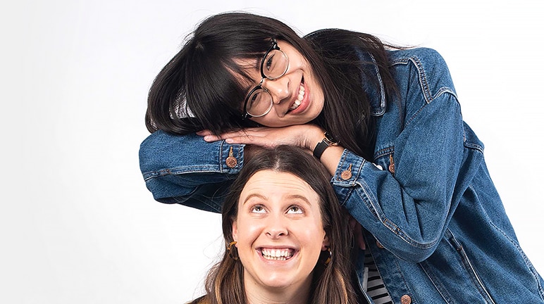 Deborah Ho rests her arms and head on Annabel Blake's head, who is seated. Both women are smiling.