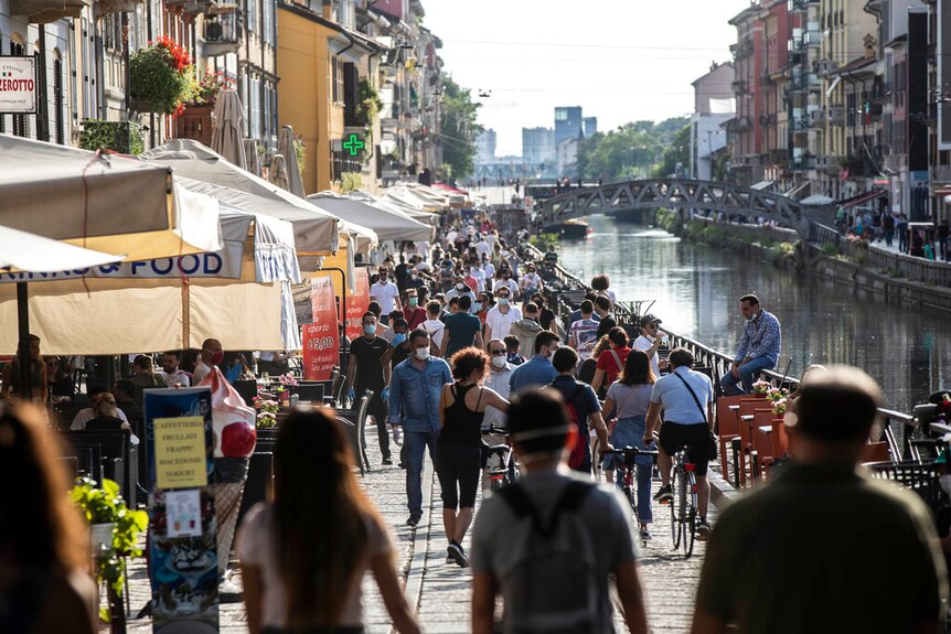 You view a large crowd of people walking along a cobblestone-lined path adjacent to a canal.