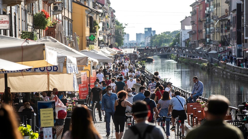 You view a large crowd of people walking along a cobblestone-lined path adjacent to a canal.