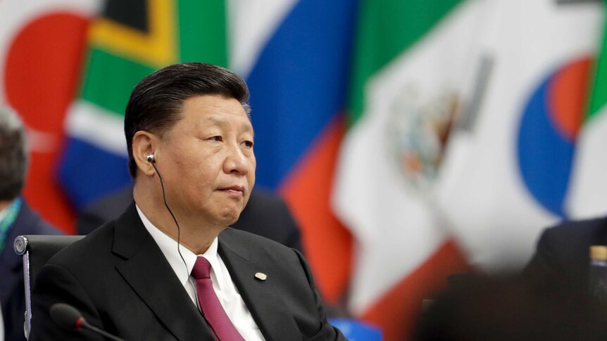 Xi Jinping, sits with an earpiece, with flags in the background.