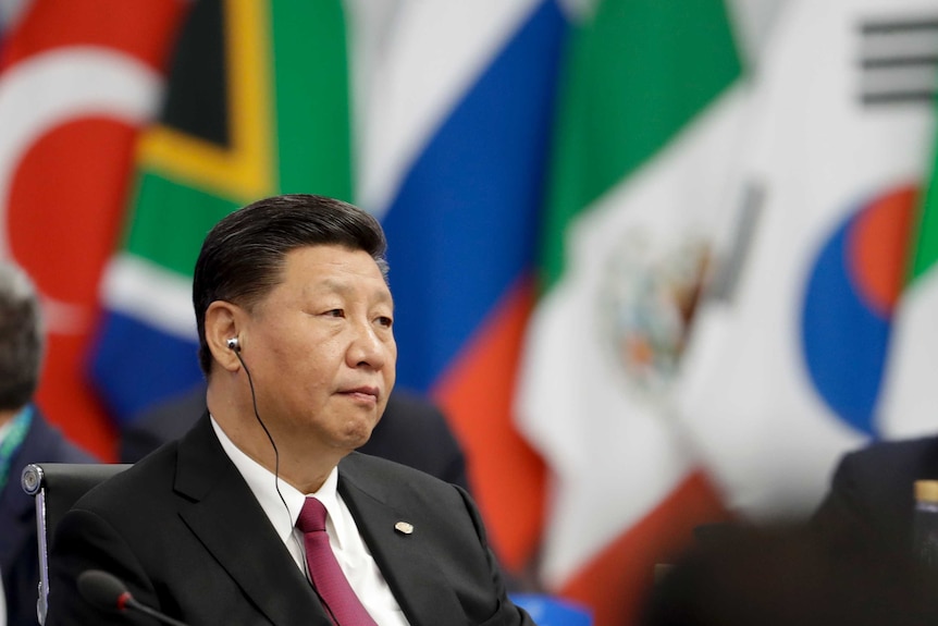 Xi Jinping, sits with an earpiece, with flags in the background.