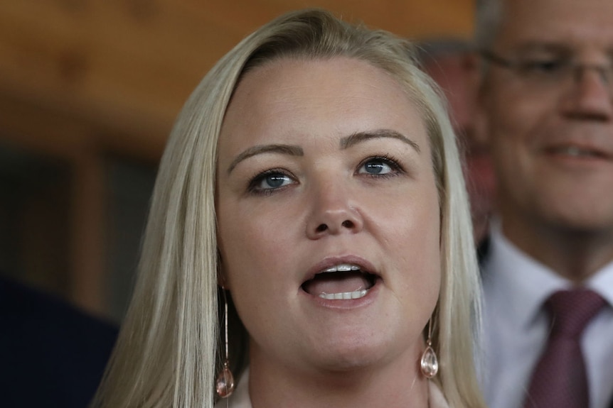 A woman with blonde hair, wearing a beige blazer, speaks to reporters. Scott Morrison stands behind her