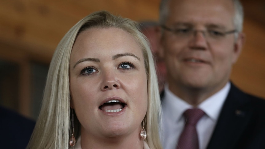 A woman with blonde hair, wearing a beige blazer, speaks to reporters. Scott Morrison stands behind her