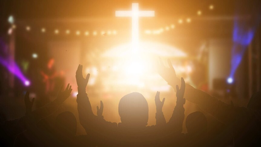 A crucifix in glowing light on stage, with Christian worshippers raising their hands in praise, at a night concert.
