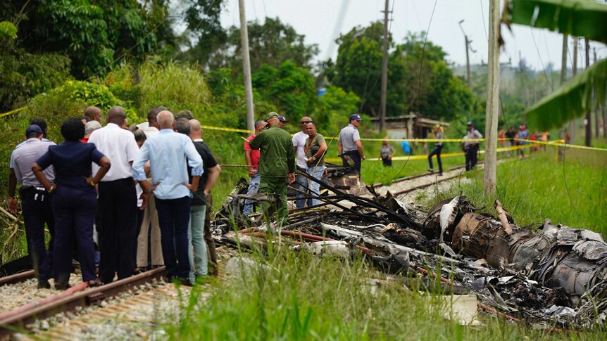 Search and rescue workers stand on train tracks next to a large piece of wreckage in a lush green field, with trees