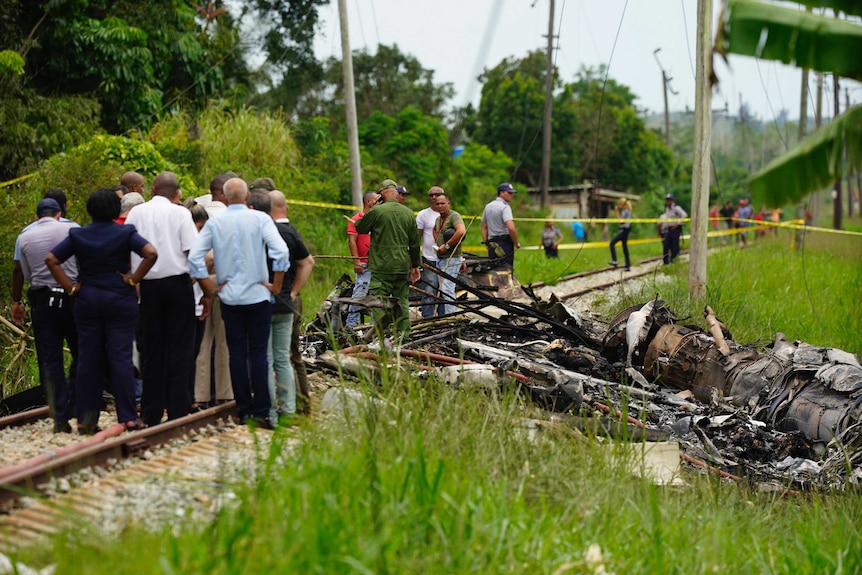 Search and rescue workers stand on train tracks next to a large piece of wreckage in a lush green field, with trees