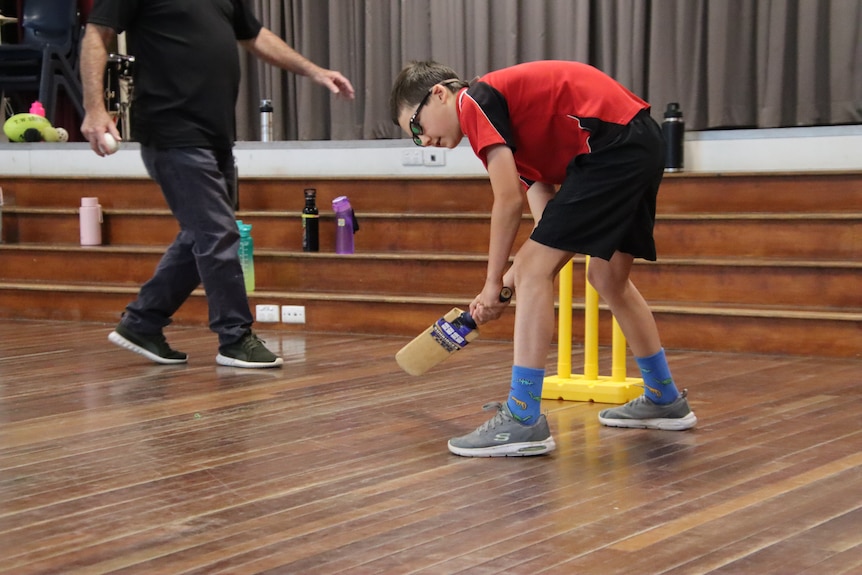 A boy wearing a red school uniform and obscuring glasses holding a cricket bat in front of a wicket in a gym