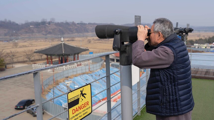 A man holds binoculars looking across a border wall.