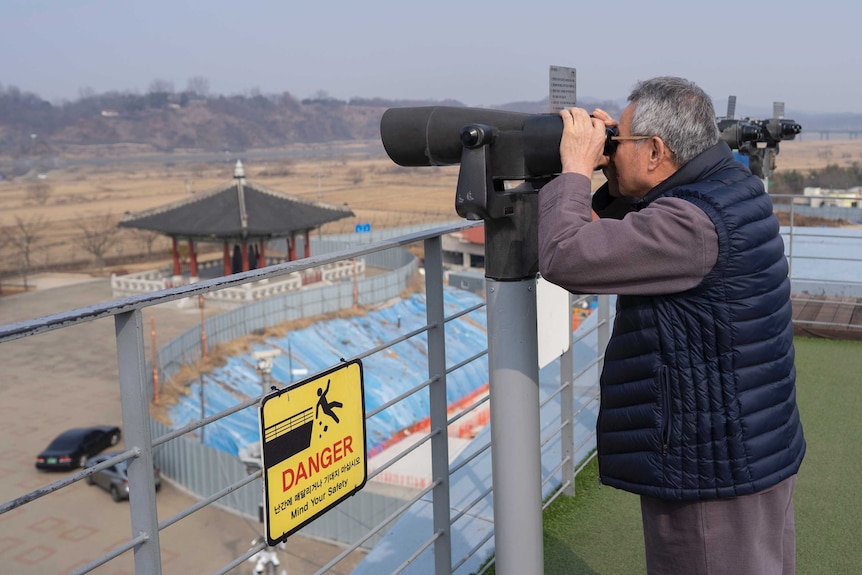 A man holds binoculars looking across a border wall.