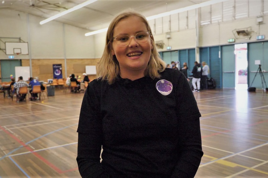 Young girl with short blond hair and glasses smiling at camera with school gymnasium in background.