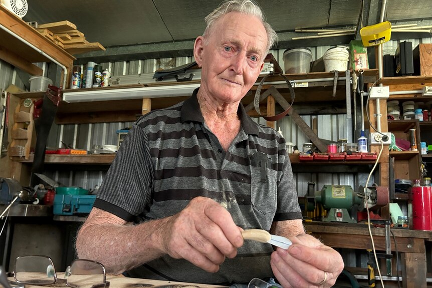 Elderly man stands in a workshop holding a small fishing lure.