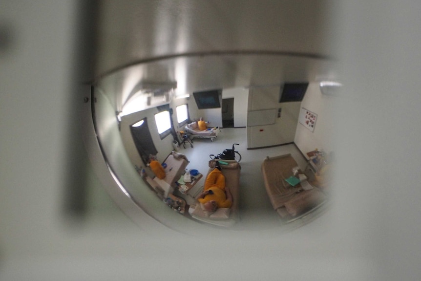 A prison mirror that reflects the image of three older prisoners lying in their beds.