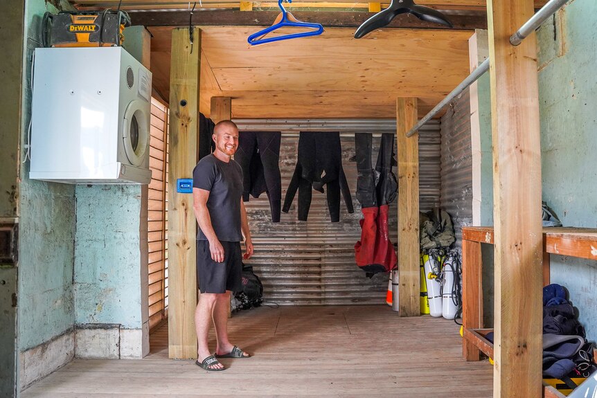 A man stands in a room with a washing machine mounted on the wall and wetsuits hanging from the ceiling.