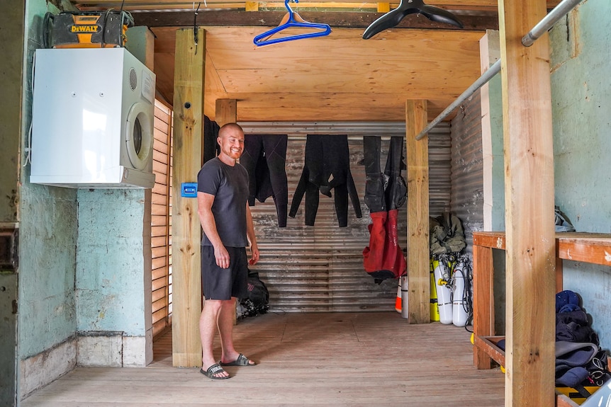 A man stands in a small shed with wetsuits hanging to dry in the background.