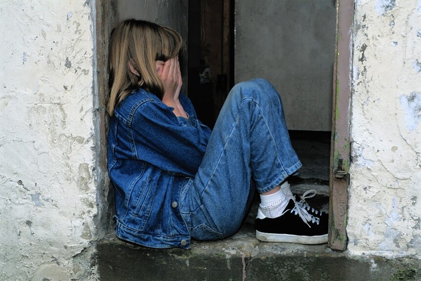 A sad child in denim jeans and jacket crouches with their hands over their face.