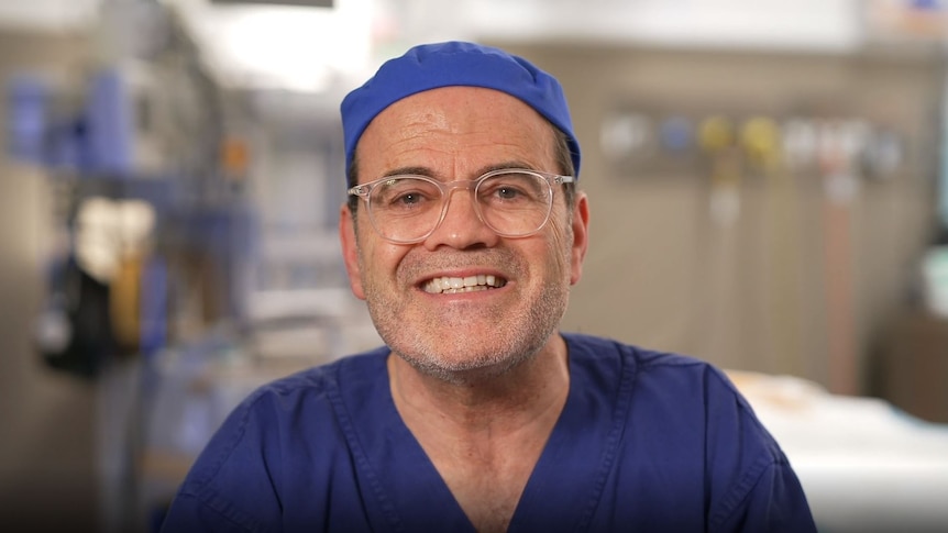 Daniel Lanzer, wearing a surgeon's scrubs, smiles, looking at the camera. He appears to be sitting in a surgery.