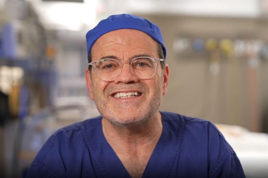 Daniel Lanzer, wearing a surgeon's scrubs, smiles, looking at the camera. He appears to be sitting in a surgery.