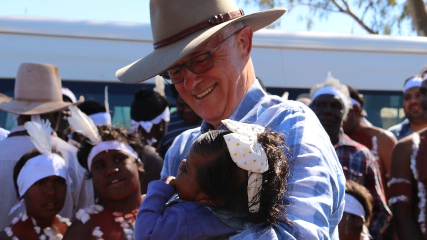 Malcolm Turnbull, wearing an Akubra, hugs a small child wearing a bow in her hair. Aboriginal performers watch in the background