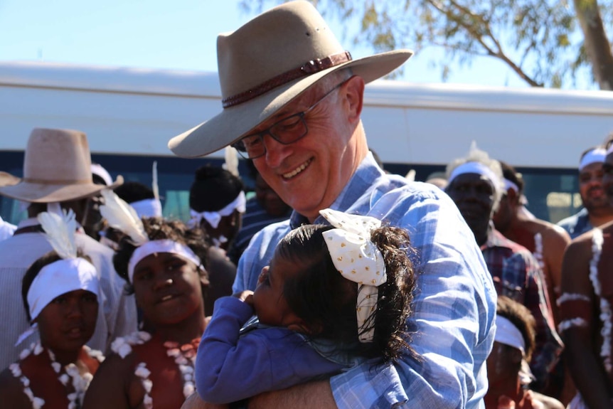 Malcolm Turnbull, wearing an Akubra, hugs a small child wearing a bow in her hair. Aboriginal performers watch in the background