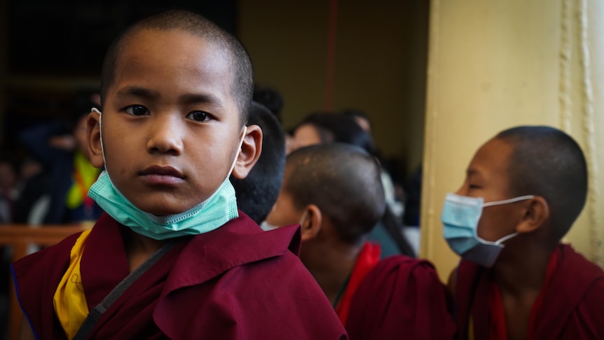 A young boy in maroon robes with a surgical mask around his chin looks into camera, kids behind him look away