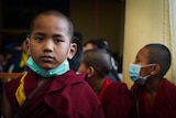 A young boy in maroon robes with a surgical mask around his chin looks into camera, kids behind him look away