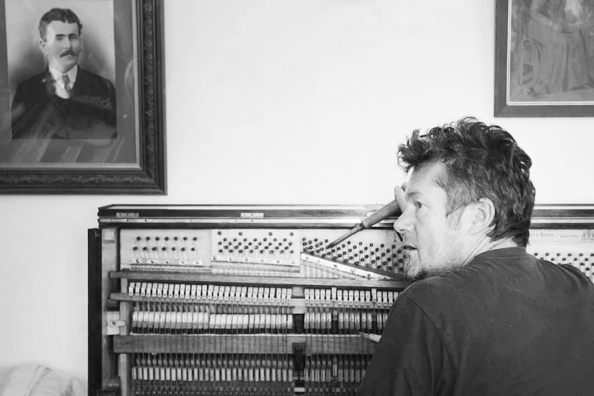 Black & white candid shot from behind of man tuning a piano looking to left. Old portraits on wall