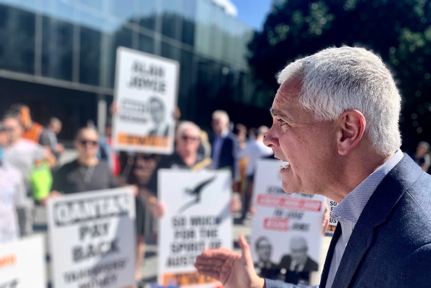 A profile shot of a man addressing a crowd, with workers holding protest signs in the background.