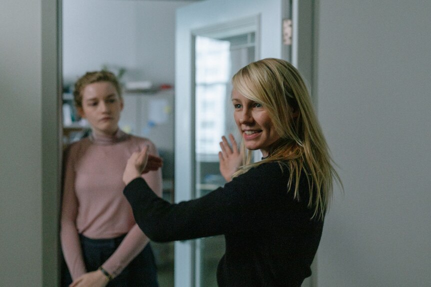 A blonde woman wears black long sleeve top, looks to left and gestures with hands as younger woman stands in doorway and watches