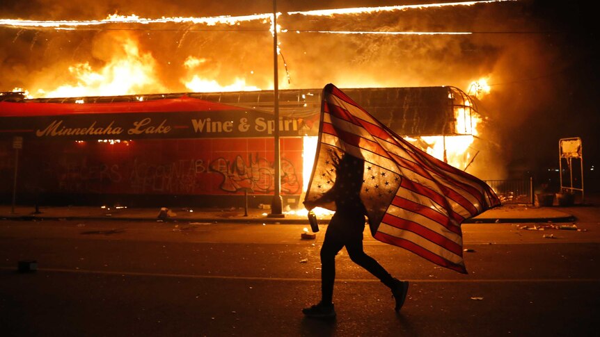 A protester carries an upside down US flag as they walk past a burning building on an empty street.