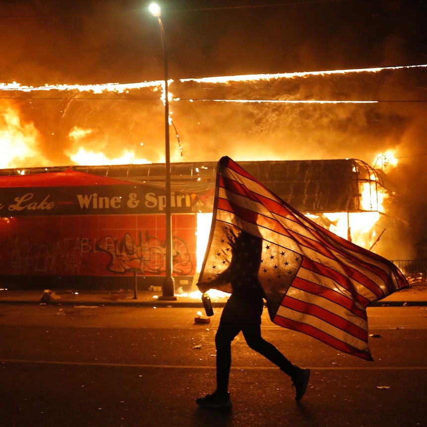 A protester carries an upside down US flag as they walk past a burning building on an empty street.
