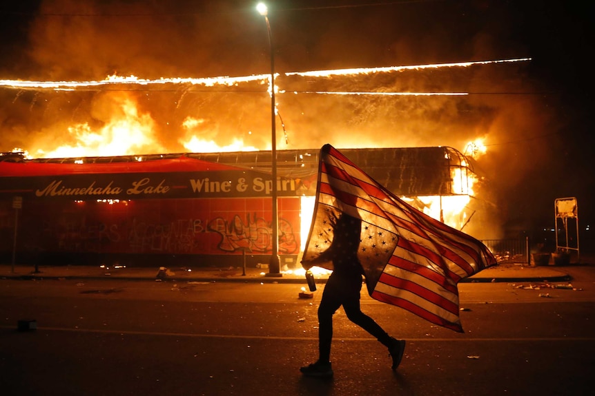 A protester carries an upside down US flag as they walk past a burning building on an empty street.