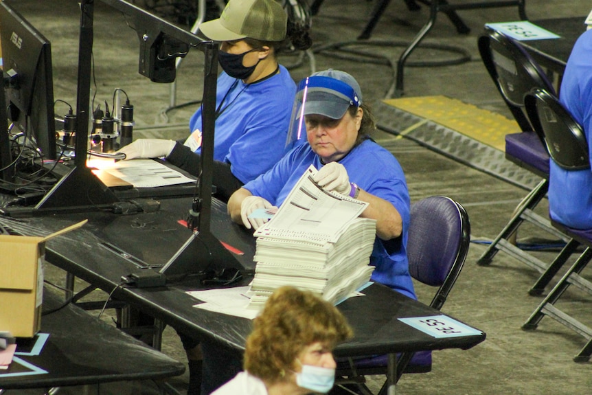 A woman in a blue shirt with a face shield grabs a ballot to place under a camera lens 