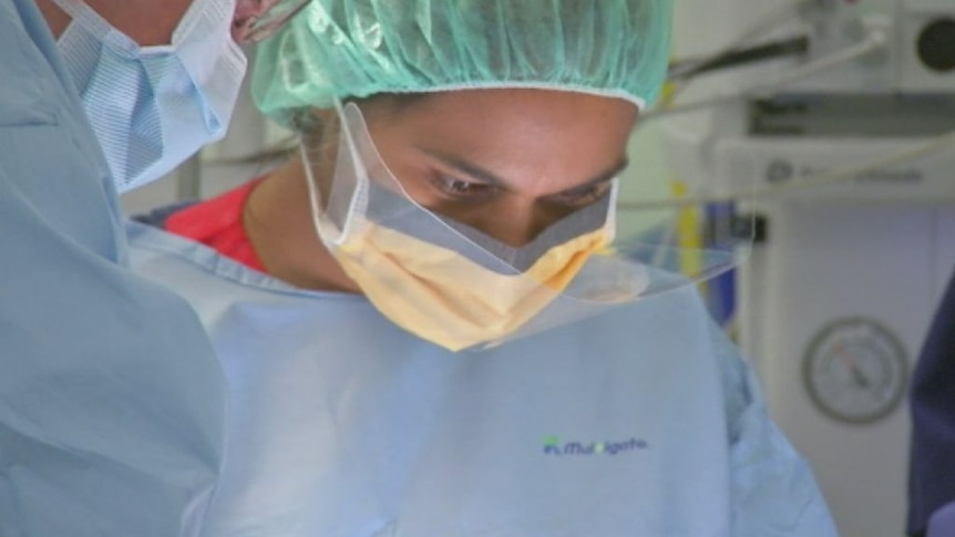 Woman in nurse scrubs and hair net with mask looks down at a patient