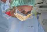 Woman in nurse scrubs and hair net with mask looks down at a patient