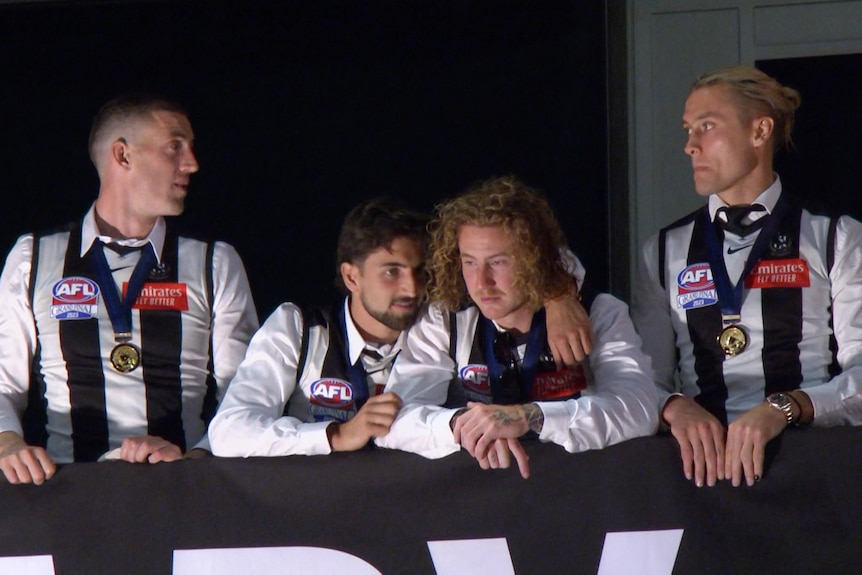 Four players wearing their Collingwood guernseys and medals stand on a podium, one with his arm around another.