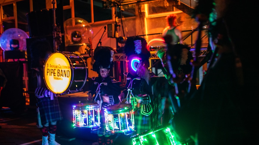 Interior night-time shot of pipe band in uniform with LED-lit drum kits, performing in club environment.