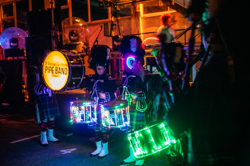 Interior night-time shot of pipe band in uniform with LED-lit drum kits, performing in club environment.