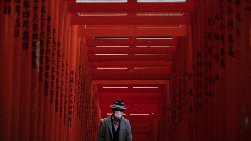 An old Japanese man walking through an orange wooden shrine with a surgical mask on his face