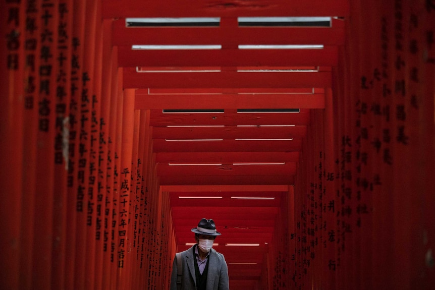 An old Japanese man walking through an orange wooden shrine with a surgical mask on his face