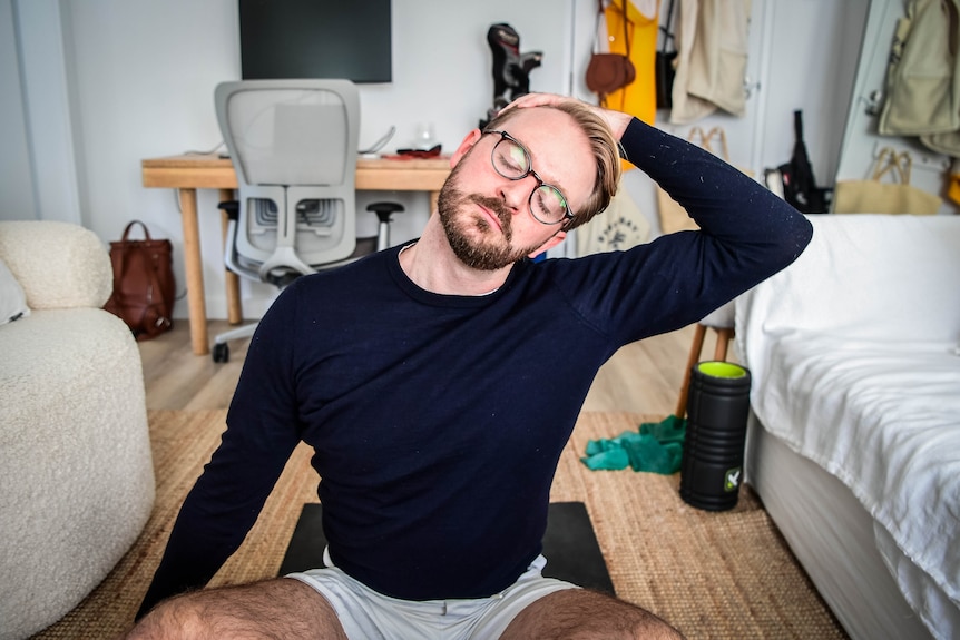 Giles sits cross-legged on the floor of his apartment. He uses one hand to push his head towards his shoulder.