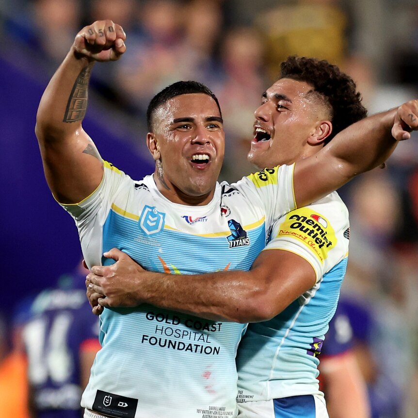 Two Gold Coast Titans players celebrate. One raising his arms celebrating while being hugged by his teammate