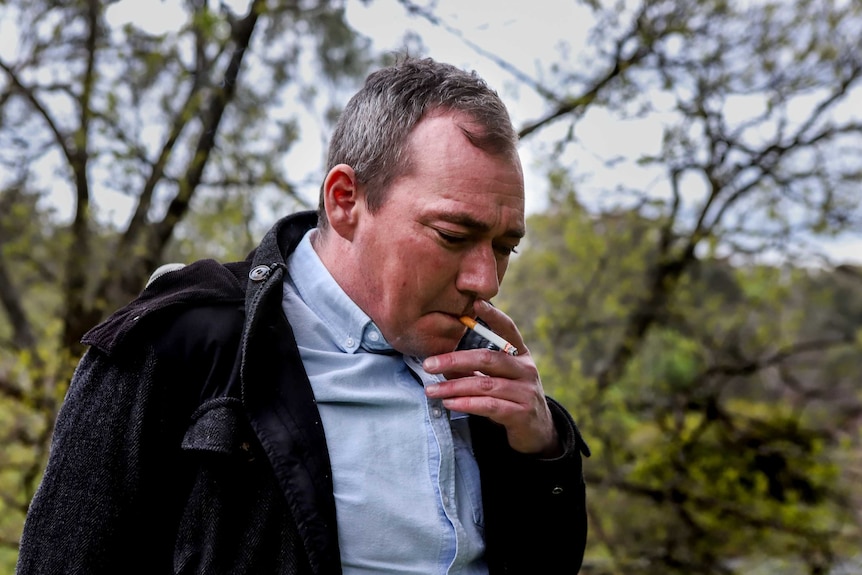 Man wearing blue collared shirt and black hooded jacket smoking a cigarette with green trees in background