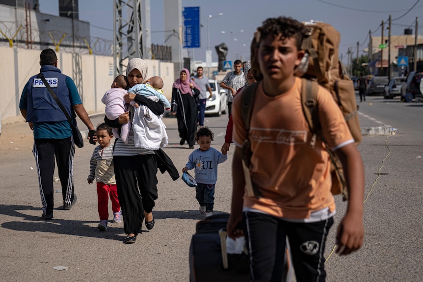 A woman walks holding two small babies in her arms as a man walks pulling a suitcase. 