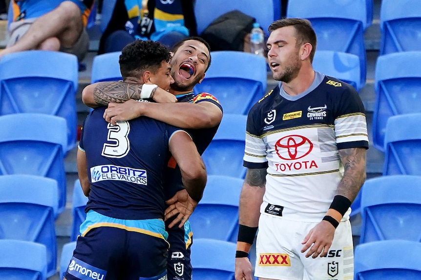 Two Gold Coast Titans NRL players embrace as they celebrate a try, as a North Queensland Cowboys opponent walks by.