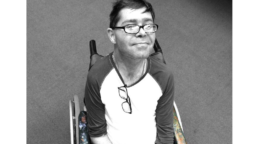 Black and white bird's eye view of a man in a wheelchair. He's quietly smiling. Paint splats on chair's arm are in colour.