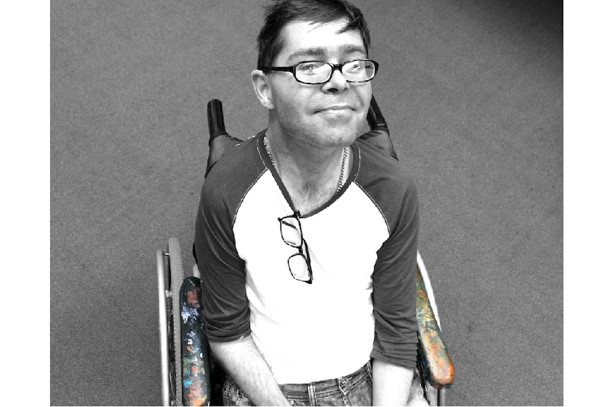 Black and white bird's eye view of a man in a wheelchair. He's quietly smiling. Paint splats on chair's arm are in colour.